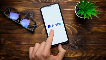 Eine Hand zeigt auf ein Smartphone, das das PayPal-Logo anzeigt, mit einer Brille und einer kleinen Pflanze in der Nähe.