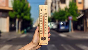 Eine Hand hält ein Holzthermometer, das hohe Temperaturen in Celsius und Fahrenheit auf einer sonnigen Straße zeigt.