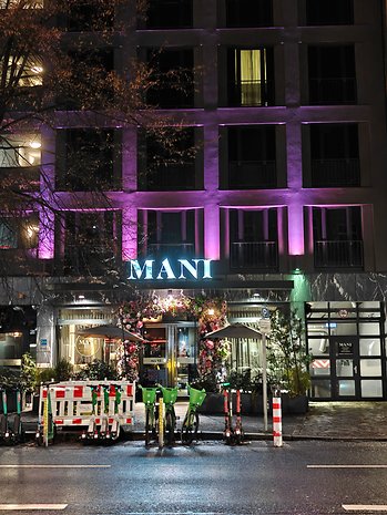 Vue nocturne d'un restaurant nommé 'MANI' avec un éclairage coloré et des vélos et trottinettes garés.