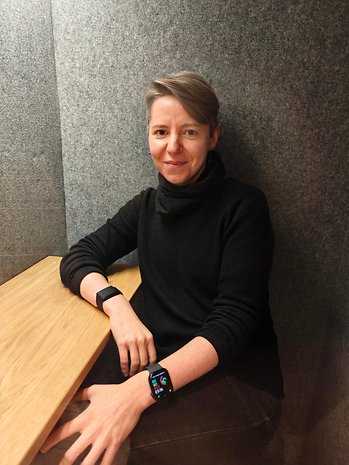 Une femme aux cheveux courts est assise à une table, portant un col roulé noir et une montre connectée.