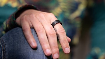 Eine Nahaufnahme einer Hand, die einen schwarzen Oura-Ring am Finger trägt und auf einem Knie ruht.