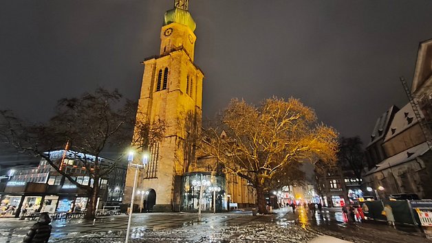Night view of a tall clock tower with an illuminated tree and people walking on a snowy square.