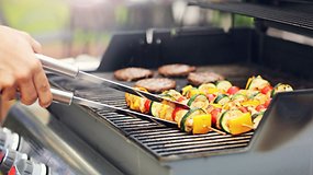 Eine Person grillt bunte Gemüsespieße und Burger auf einem Grill.